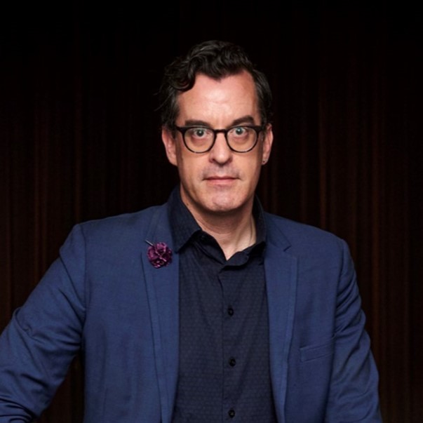 A man wearing a blue suit with a navy blue shirt in front of a black backdrop. He is wearing glasses and has a purple flower pinned on the right side of his suit.